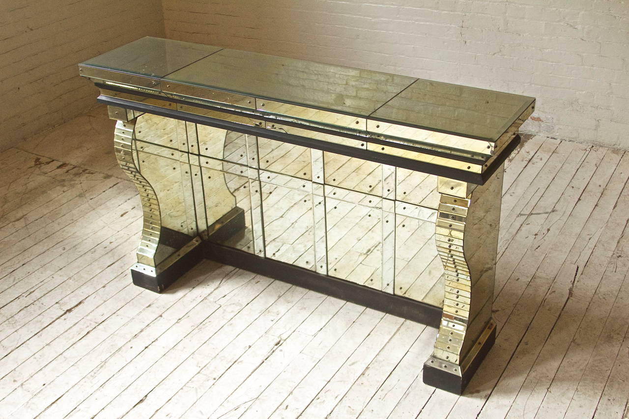 Stunning neoclassical style wall console in hand-cut glass with three panel top supported by corbel legs. Mirrored back panel, ebonized wood base, natural patina.