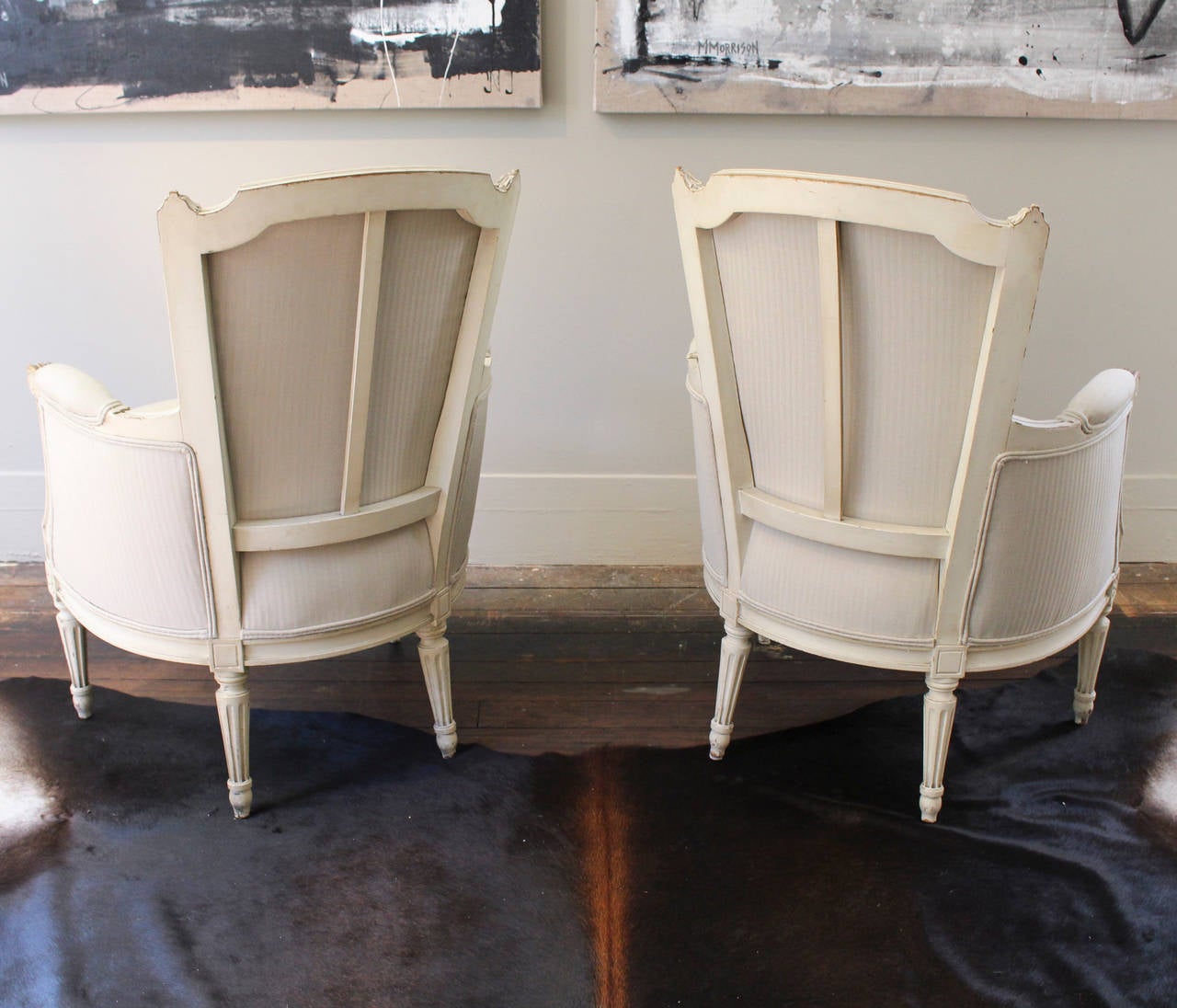 Pair of bergeres, 19th century France, in time worn oyster paint raised on rosette-topped tapering fluted legs. Newly upholstered in a natural shadow-striped Rogers & Goffigon fabric, the chairs feature padded elbow rests on scroll-curved arms.