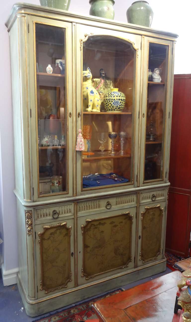 A Louis XVI style celedon painted and giltwood lighted vitrine with three beveled glass doors above three carved and fluted drawers with bronze pulls over three cabinets. Shelves are adjustable. The door panels are painted with a Chinese design.