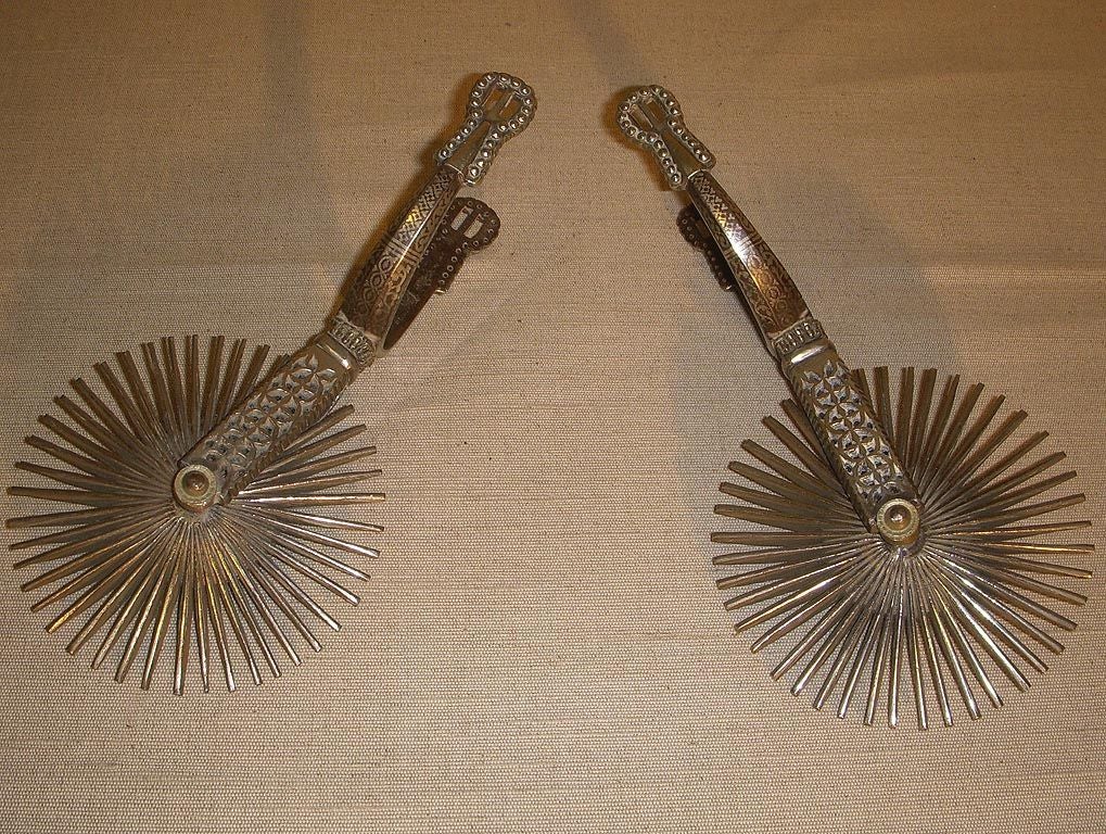 Distinctive Pair Silver Metal Rowelled Spurs Ornamented with an Incised Geometric Design and Rotating Toothed Wheels. Latin America, Late 19th / Early 20th Century.<br />
<br />
10 inches long / 4.8 inches diameter of round spurs<br />
<br
