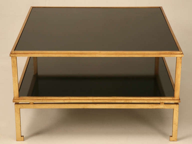 Mesa de centro de inspiración francesa moderna de mediados de siglo con un acabado dorado aplicado a mano a la antigua usanza. Se muestra con nuestra configuración estándar de tapa de cristal negro y estante inferior, aunque nuestra Mesa de centro