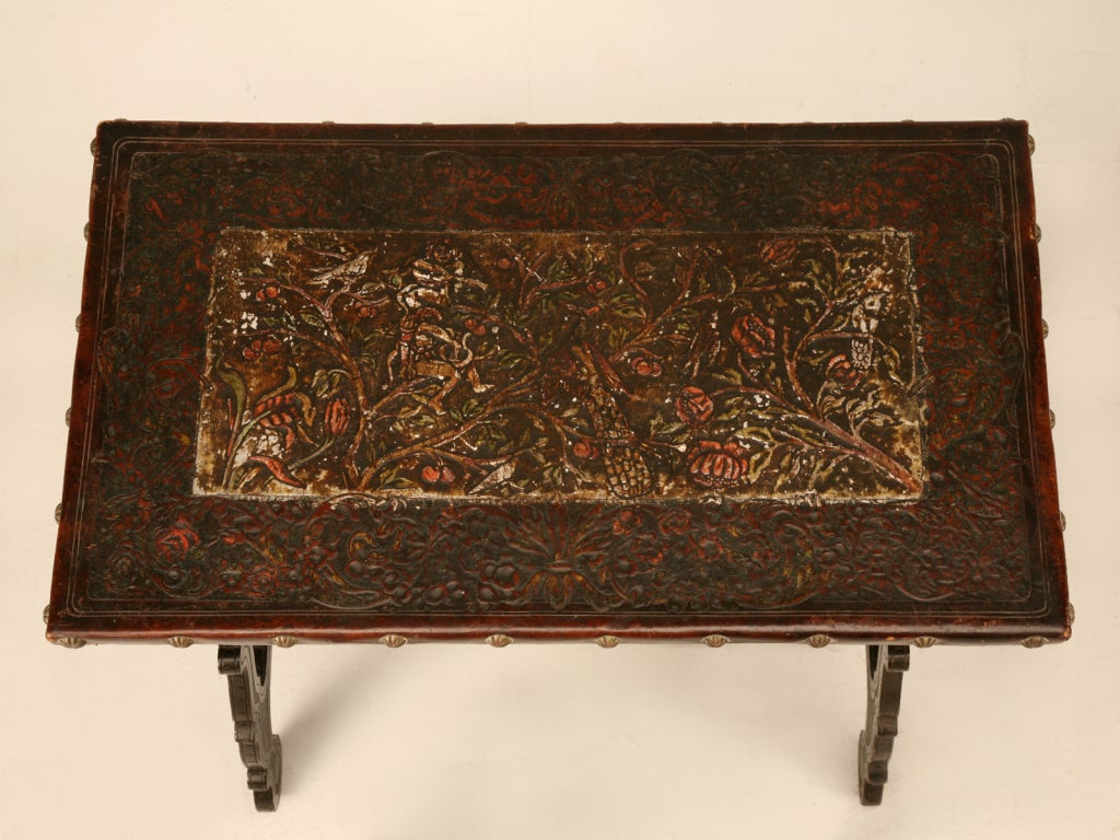 This spectacular petite antique Spanish table with its heavily carved solid oak legs and frame which are steadied by a stunning hand-wrought iron stretcher and topped with one of the most phenomenal original tooled and painted leather tops we've