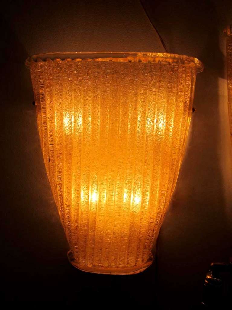 Pair of amber color Murano glass sconces with ripple texture. The sconces have brass hardware and back plates.  Each sconce has four sockets inside. Wired for US standards. We have three pair available.
