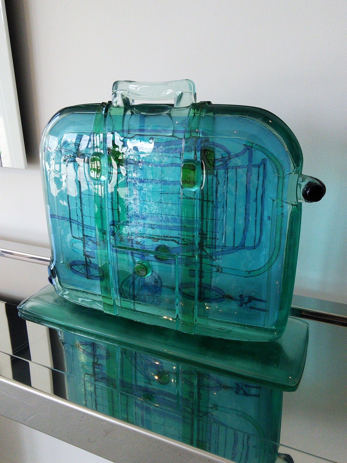 Green cast glass sculpture in the shape of a suitcase. The colors of the glass range in various hues from green, sea-foam green to blue. The interior of the glass there are outlined silhouettes of the contents of the suitcase -- a shopping cart.