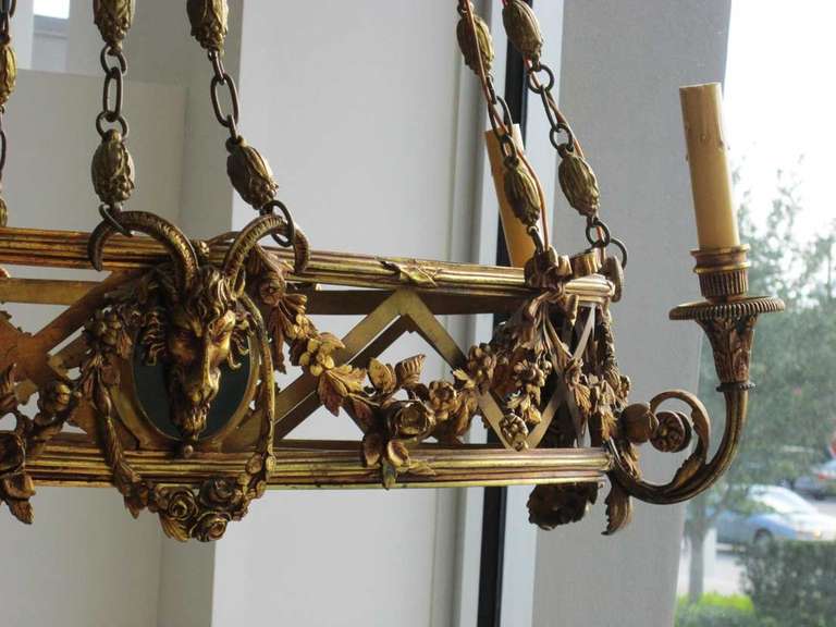 An oval bronze chandelier  Revivalist in style - featuring dramatic ram heads and garland details strung over  diamond patterned lattice work.The fixture has four faux candle stick sockets that are mounted on gilt scroll shaped pieces of garland