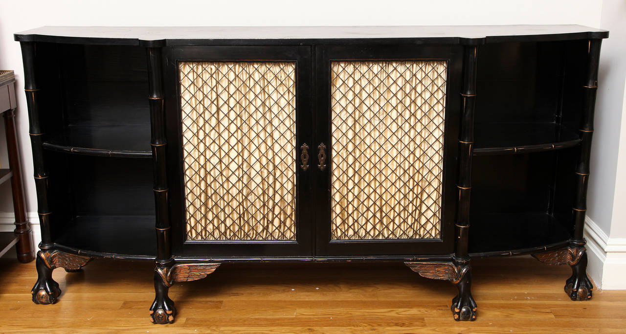 An English Regency style bow front long sideboard. The pair of wire mesh cabinet doors having shirred fabric concealing interior fitted with shelves, flanked on each side by open shelves framed with vertical sections of faux bamboo. The base