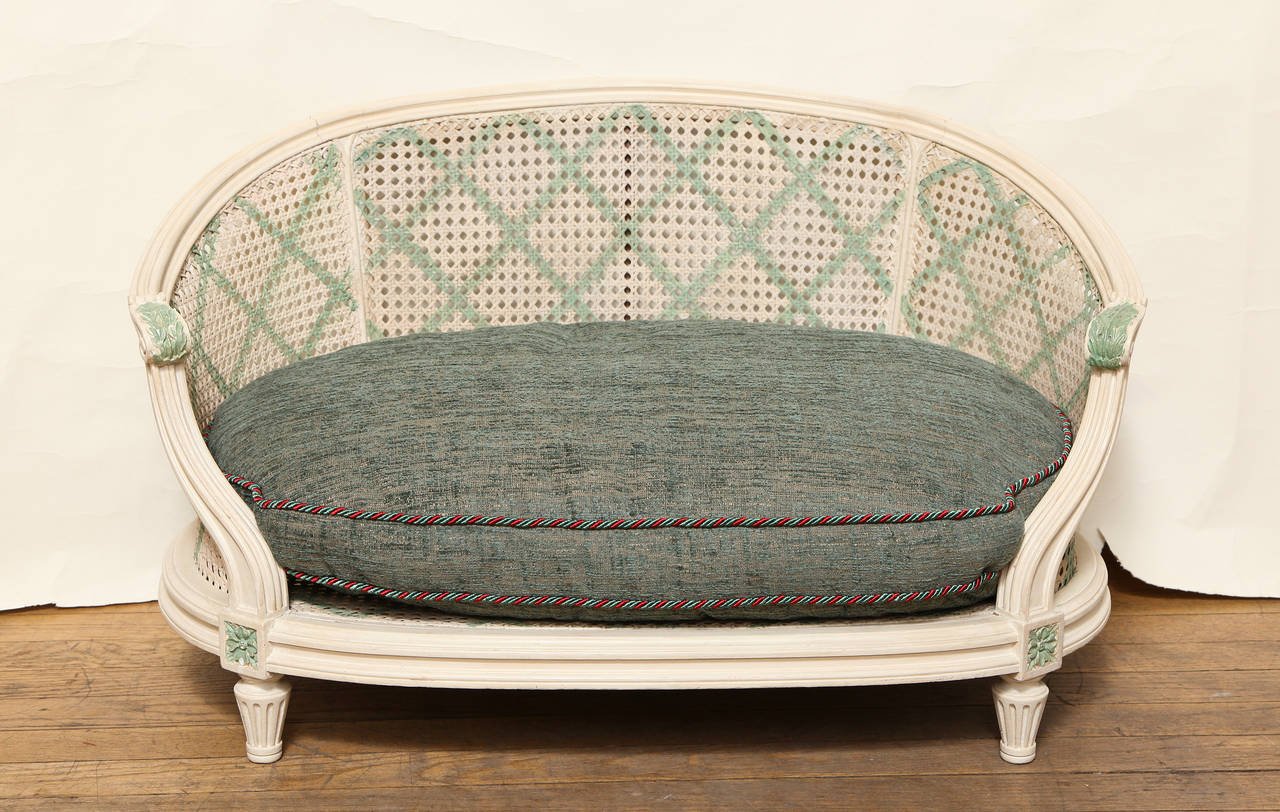 A French Louis XVI style white painted caned dog bed. The arms with leafed shaped detail, the arched back, sides, and base with contrasting green striping. The back and sides with trellis patterned stenciling. The frame supported short round
