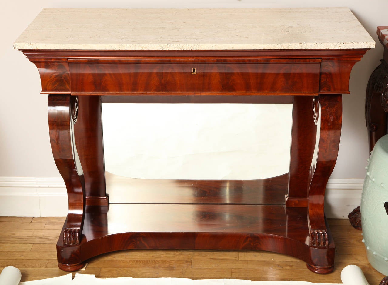 A French Charles X period mahogany console with drawer in crotch mahogany veneered apron, supported by curved bracket legs backed by mirror. The lions paw feet resting on plinth and having travertine stone top (replaced).