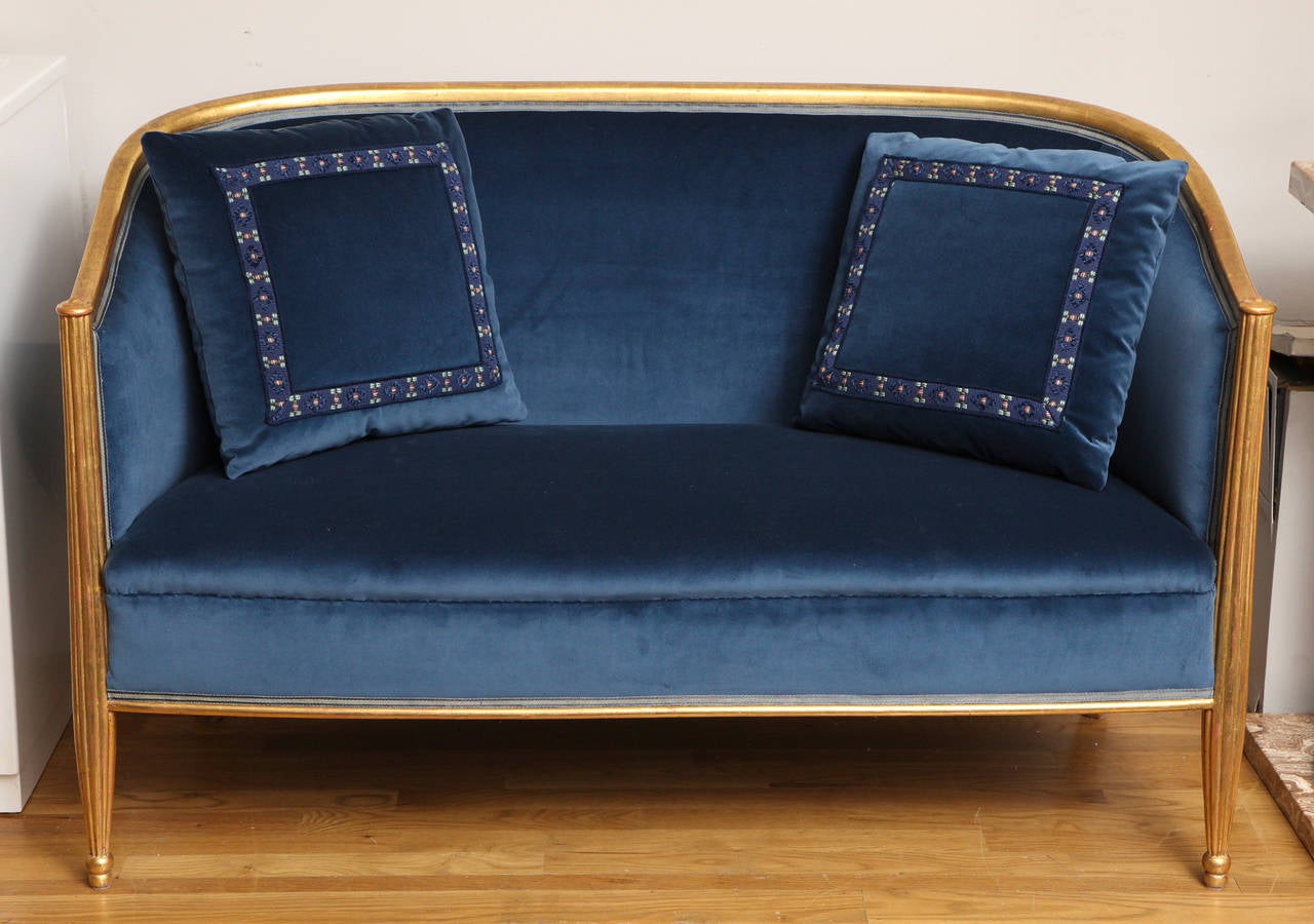 A French Art Deco sofa with carved in giltwood frame. The curved back joined to base by reeded tapering vertical elements forming front leg, the back supported by splayed sabre-form legs. Sofa newly upholstered in blue cotton velvet with matching