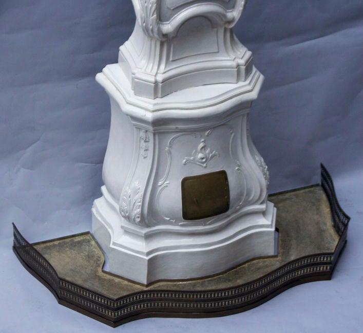 An Italian Rococo porcelain terra cotta stove with the lower section having Bombay front and sides and hinged brass door to facilitate use as stove. Upper section with bell flower motif below Rococo cartouche, the top surmounted by finial. Lower