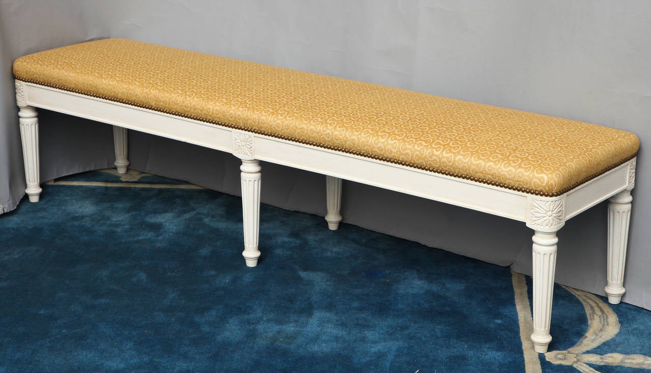 A rectangular bench with upholstered seat. The new upholstery trimmed with nailheads above apron with rounded corners having hand carved corner rosettes above round tapering fluted legs. The frame, molded edges, rosettes, and fluting having antiqued