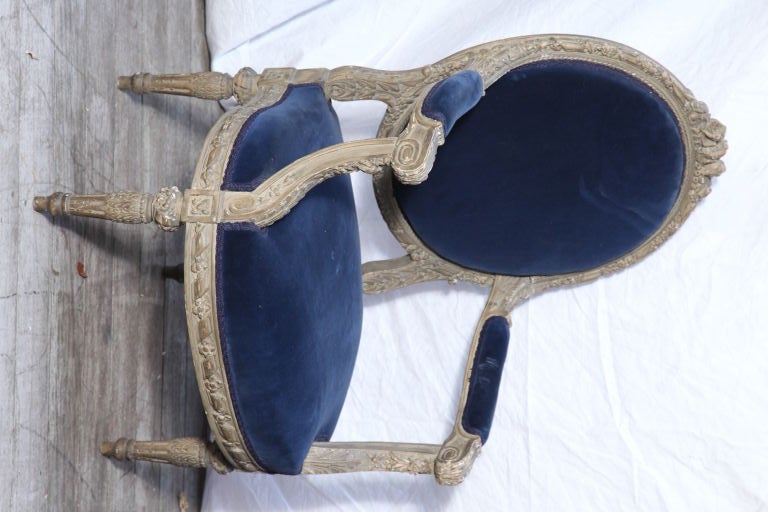 A pair of French Louis XVI style open arm chairs with carved beechwood frames painted gray, the leaf and floral motif detailed frames having tapering fluted legs and ribbon carving.  Upholstered seats, back, and armrests covered in blue velvet.