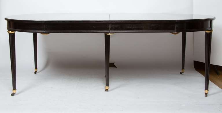 A French Louis XVI-style ebonized racetrack oval dining table, each end supported by round, tapering fluted legs terminating in bronze sabots with casters, the tabletop opening to accommodate four 22” extension leaves which can be used in any