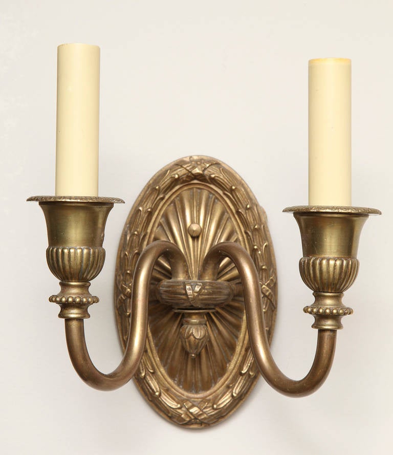 A pair of French Louis XVI-style two-light wall sconces with oval backplate having laurel and cross band border issuing candle arms with vase shaped bobeches. Backplate measures: 6.25