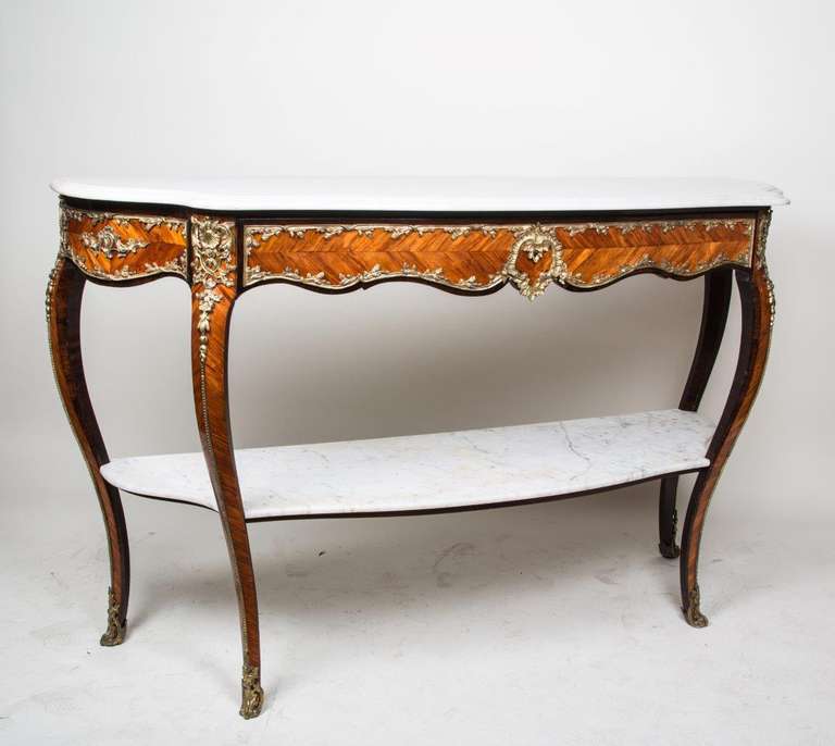 A two tiered French Louis XV style console with tulip wood veneered carcass having contrasting rosewood edging and mounted with fine leaf motif, bronze mounts surrounding drawer in upper apron, and having two carrera marble surfaces. The upper