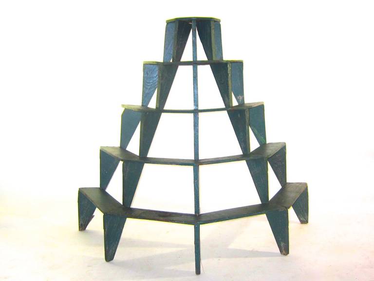 A handmade painted  five tiered wood plant stand.  The  custom design lends a delicate feeling to the structure which has a small older patch on one shelf . The unit shows  a gentle use  considering the purpose and age . There are areas of worn 