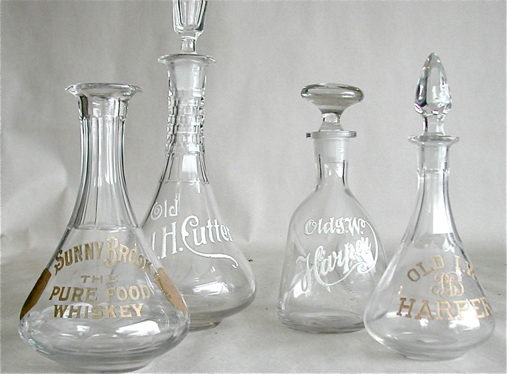 The four back bar or table top whiskey decanter bottles bear the advertising label or logo of their respective brands. Two ar embossed with porcelain enameel and two are incised with gold lettering and shield. The smaller I.W. Harper is a pinch