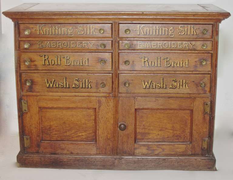 This hard to find Silk thread spool cabinet has eight drawers and a double door storage  space entry on the lower portion. It is not branded and has the unusual feature of a marble insert top. The lettering is gilded and shadowed drawer content