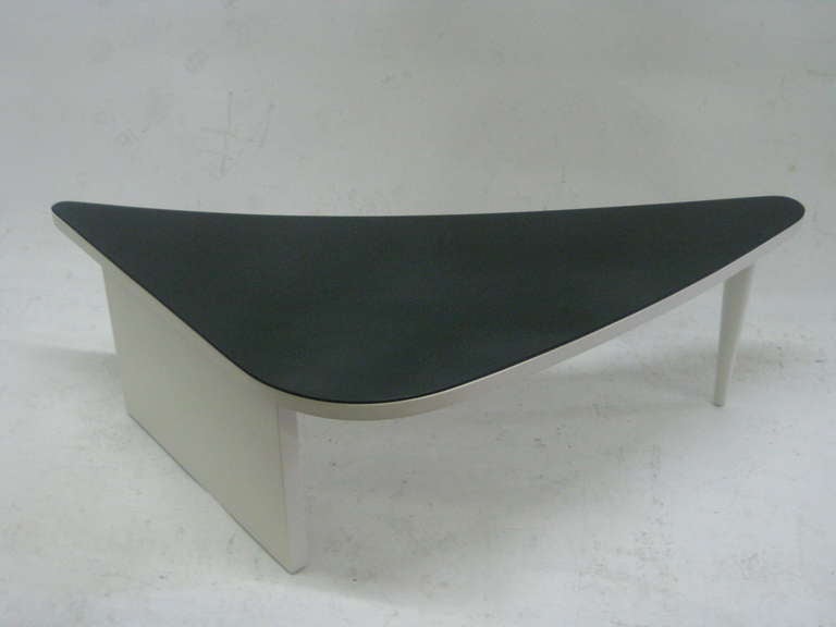 This elegant cocktail table features a triangular form with rounded edges in black glass atop white lacquer frame. Supporting the table is a solid rectilinear leg which is balanced by a tapered leg on the opposite end.