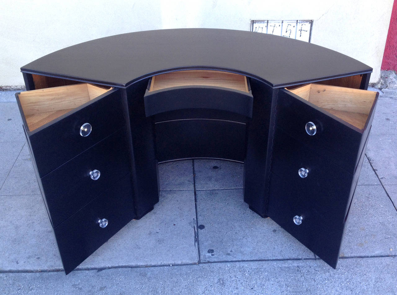 This curved half circle desk features six swing-out drawers that flank a single central pen drawer. The back of the desk has two open shelves for storage or display. This piece, recently refinished in black, has two types of Lucite knobs, which are