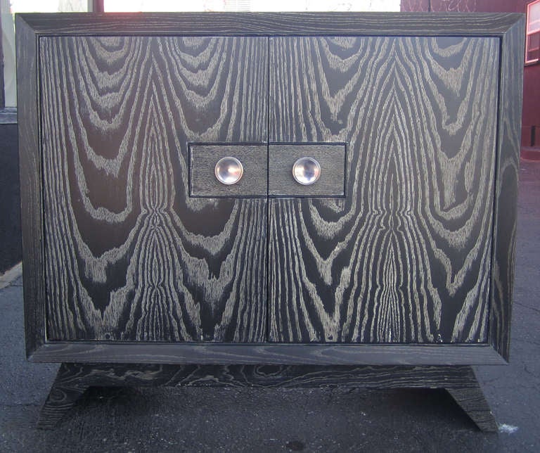 This two-door cabinet by Paul Frankl for Brown Saltman features silver-tone pulls set of by a black cerused finish. The doors open to reveal a two-tier shelf. The petite credenza rests on four splayed feet.