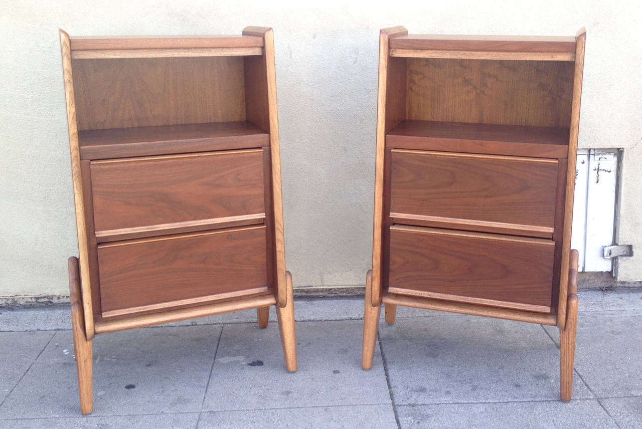 This pair of French walnut cabinets feature a very mid-century modern design with a slightly flared design and structural off-tone wooden legs. Each cabinet holds two drawers, which support an open cavity at the top.
