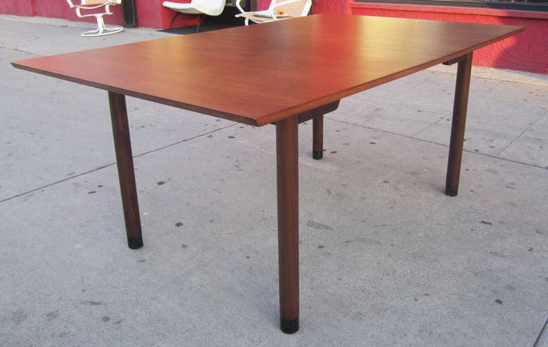This elegant dining extension table by Edward Wormley for Dunbar features  a walnut top supported by a slightly darker walnut skirt and legs. The cylindrical legs are finished with the original black leather sabots. The table is 68