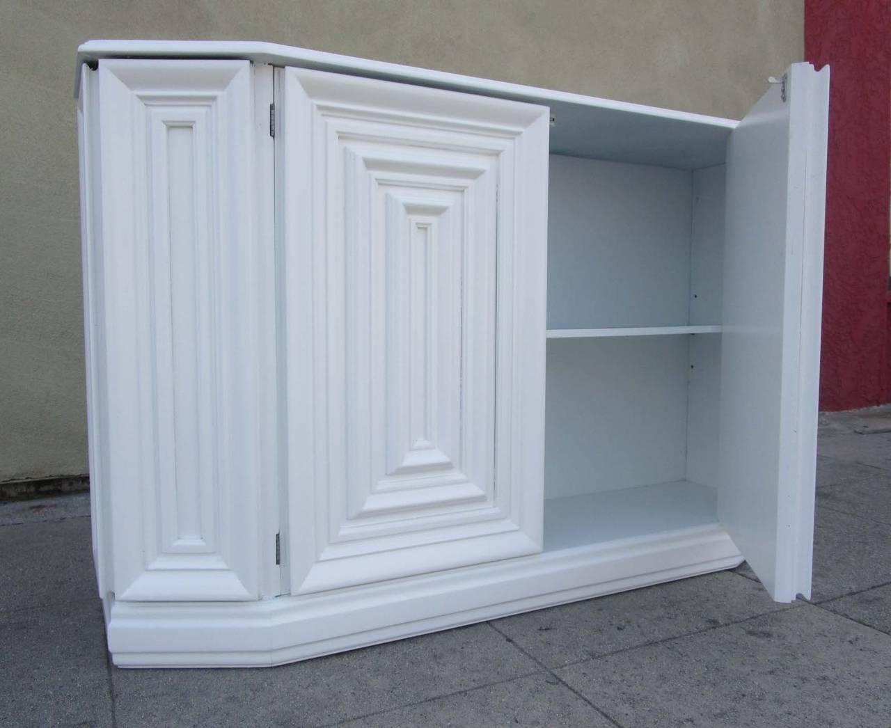 This console cabinet features two doors which hide a single shelf. The piece, made of white lacquered wood, has intricate rectangular ribbing on its front .