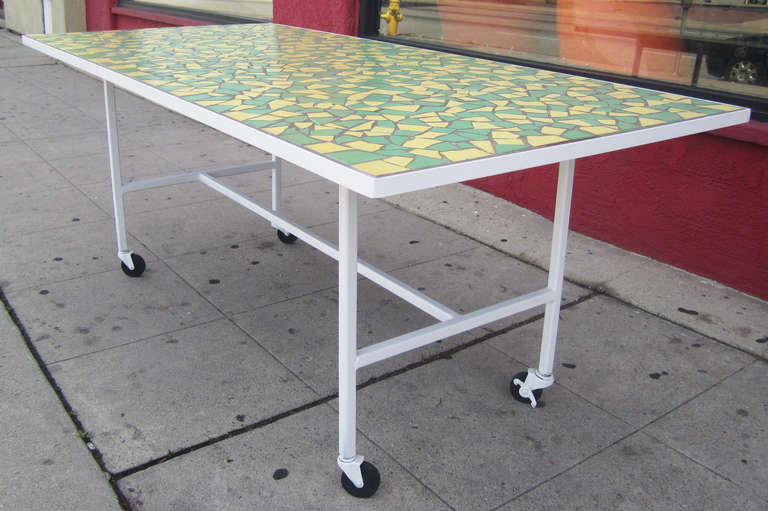 A modern dining table comprised of a green and yellow tiled mosaic top and a heavy wrought iron base, which has been recently repainted bright white to show off the summery hues of the surface. This piece, resting on casters, can be used either