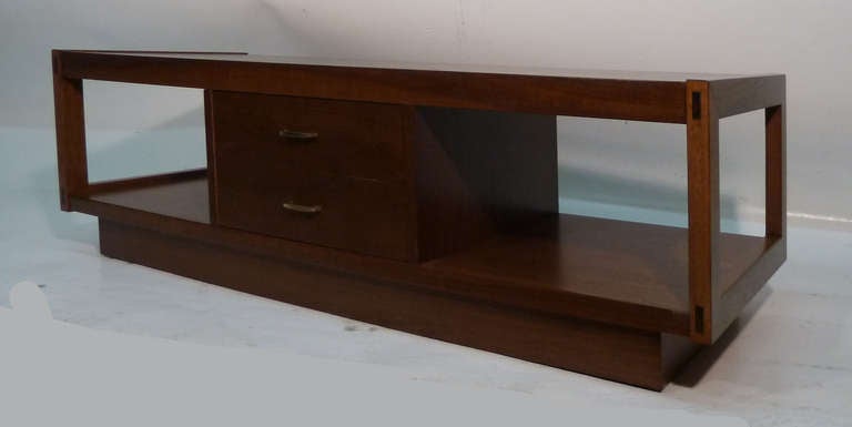 This highly structured, mid-century modern coffee table by Lane is rendered in walnut and brass and features plenty of storage including a large center drawer flanked by two open shelves ideal for the display of magazines and books. The piece's