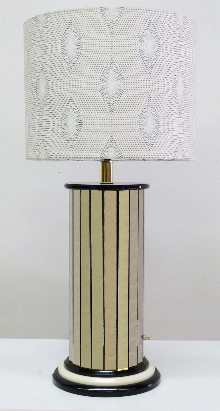 Glamorous pair of 1970s table lamps with vertical mirrored panels around a column body. Mounted on stacked ivory and black disks on a black painted wood base. Wired and in working condition. Use 100W max bulbs. New fabric shades with optical designs