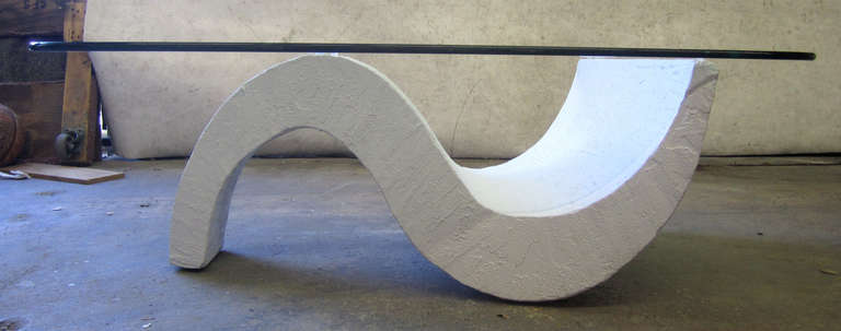 A 1980s coffee table comprised of a triangular, s-shaped base in textured concrete which supports a rectangular glass top with rounded edges.