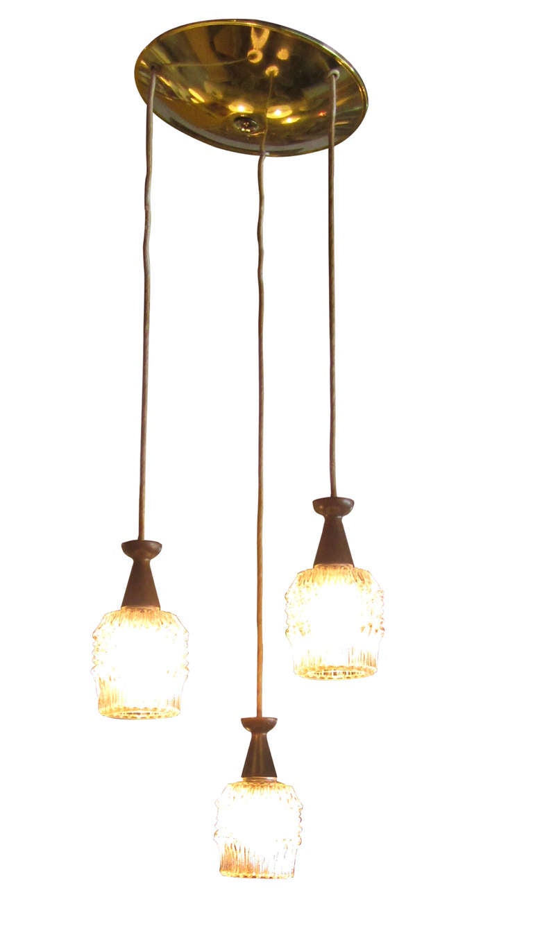 This mid-century modern lamp features three single pendants hanging at varying lengths from a disc-shaped brass canopy. Each light is comprised of a grape-motif shade in glass, a walnut pendant holder, and rope which attaches it to the brass