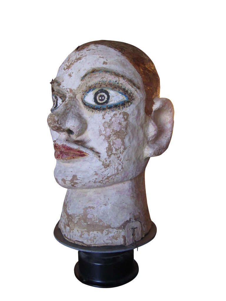 A large-scale papier mache carnival head resting atop a rotating base. The skillfully rendered head has a lively, realistic expression emphasized by hand-painted decoration and real hair used in the eyebrows and mustache.