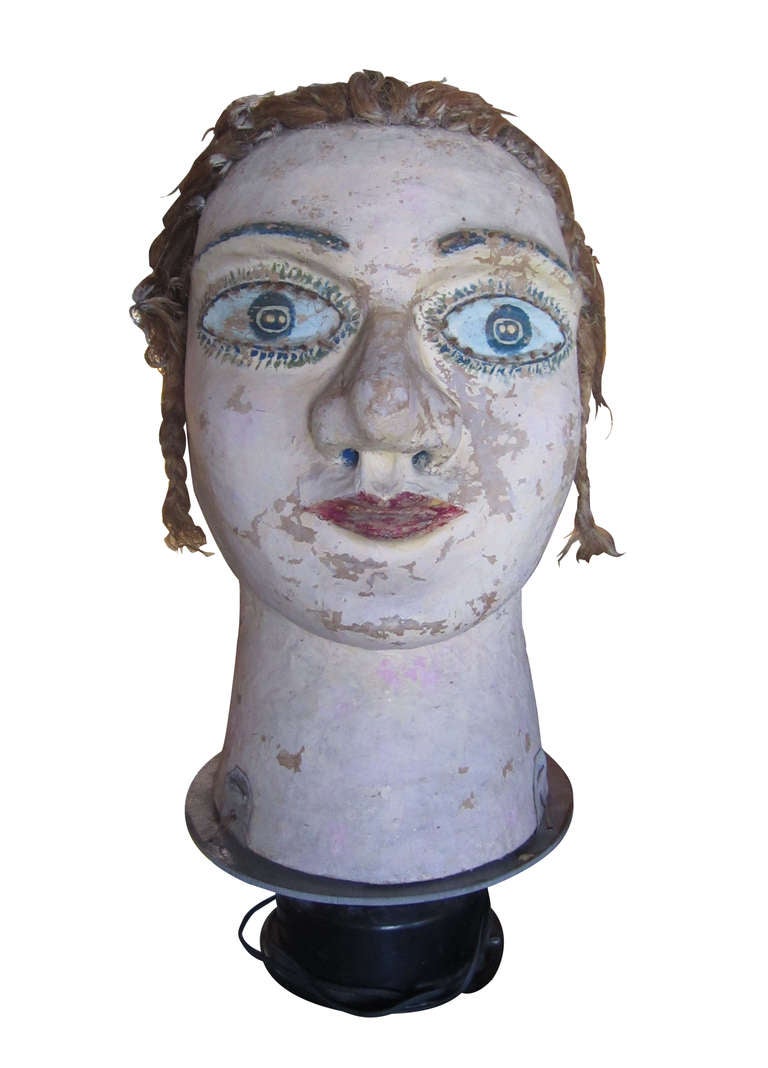A large carnival head rendered in paper mache and real hair that is tied back with a flower. The piece rests atop a rotating black plastic base.