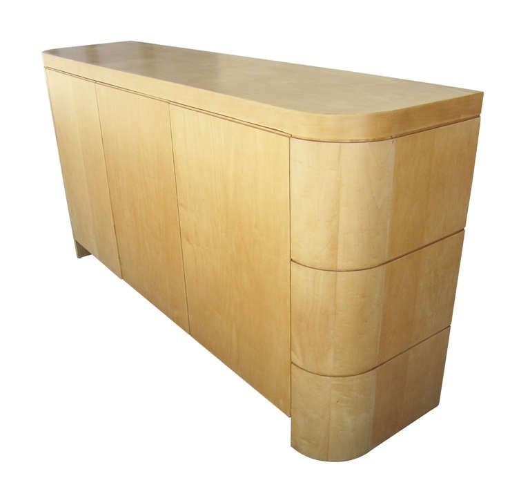 This three door credenza features an elegant, rounded shape in maple. Two doors open to reveal a large two-tiered shelf space surmounted by a drawer, which is found again as a smaller version behind the third door.