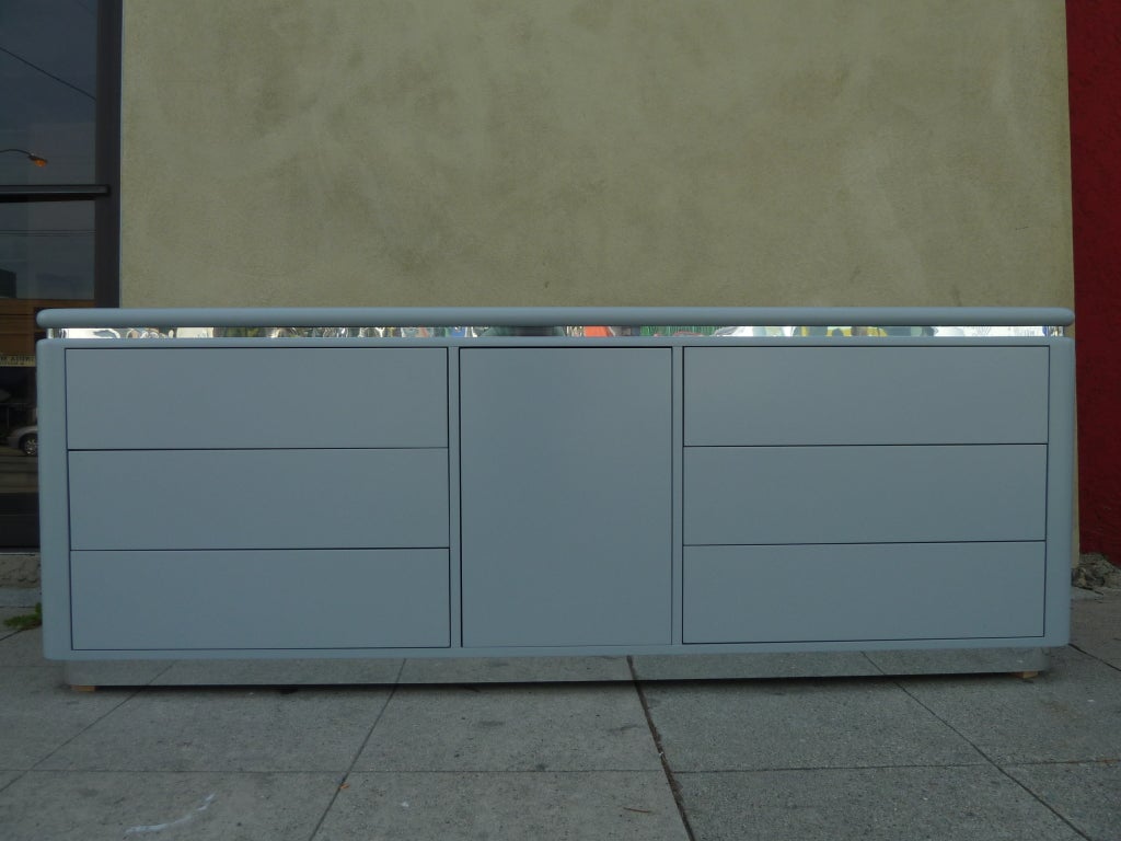 This grey lacquered chest of drawers by Lane features plenty of storage with six push-to-open drawers and a cabinet with two shelves. Inset above the drawers is a narrow chromed band which surrounds three sides. The piece is softly rounded at the