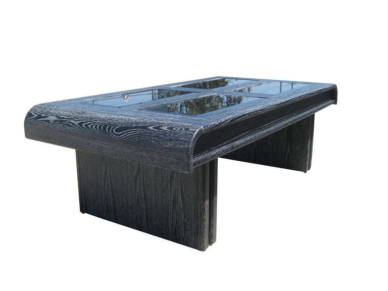 This coffee table features four smoked mirror panels which rest in a wooden base with a black and eggshell ceruse finish. The curve of the table's surface coupled with the thick, dual legs brings to mind the designs of Paul Frankl.