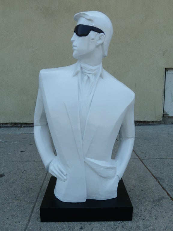 This playful bust depicts a dapper young man starring confidently over his shoulder. The man is rendered entirely in white with the striking exception of his black sunglasses. The sculpture rests on a rectangular black base.