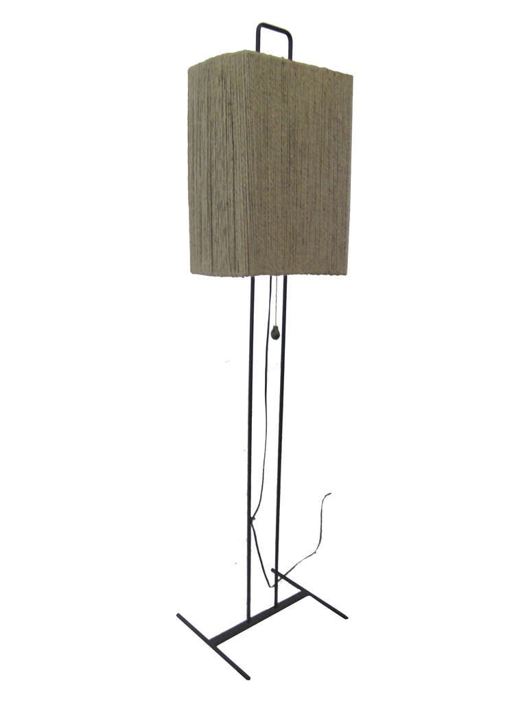 This floor lamp features a rectilinear frame in iron which supports a brown, rectangular shade. The shade consists of an iron frame which is entirely wrapped in natural brown yarn that lets light pass through when illuminated. This piece is wired