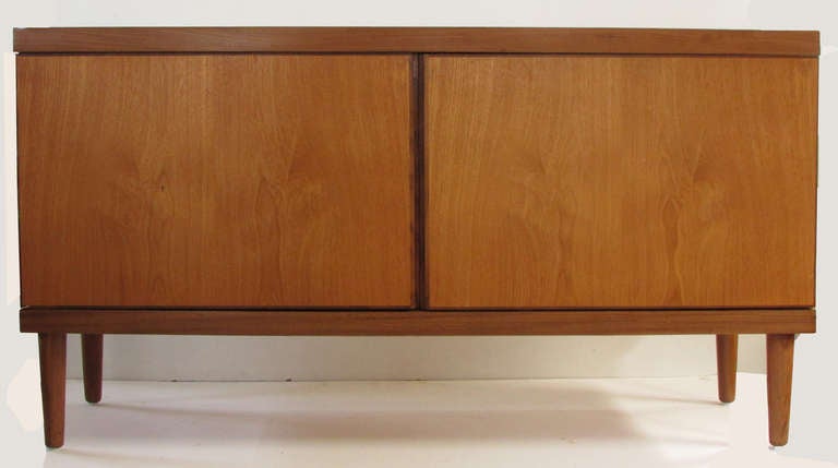 This classic, Danish modern credenza by Hans Olsen is ideal for compact settings. The piece features richly grained, amber-toned teak set off by banded contrasting edges as well as solid, tapered legs (unlike the stainless supports which were