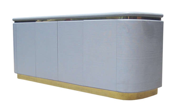 A sophisticated four-door credenza featuring a grass-cloth finish in dove grey with brass banding at the base and just under the surface. The rounded frame of this 1970s piece has four push-lock doors which open to reveal both shelf space and two