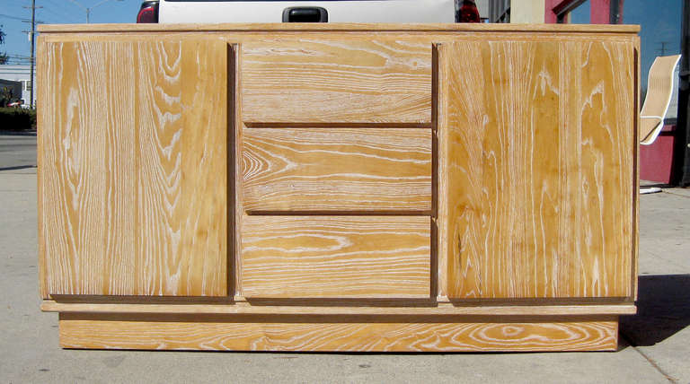 A cerused case piece in light wood featuring two doors and three pull-out drawers. The unmarked piece sits on a slightly recessed base in the same finish.