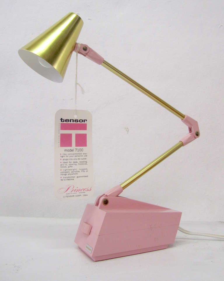 This adorable desk lamp by Tensor features a pink plastic base and joints complemented by a brass shade and arms. The lamp retains its original tags, which indicate the model, original retail price, and uses. The lamp is wired and in working