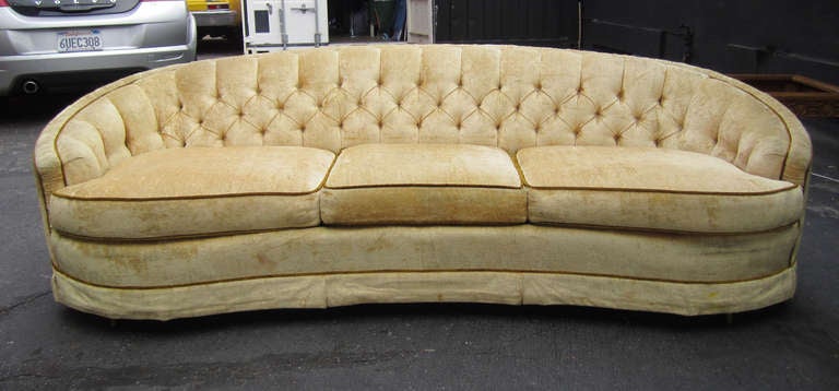 This elegant Hollywood Regency three-seat sofa features a rounded, tufted back upholstered in champagne-toned velvet and finished with olive piping. This is the original upholstery. It would stand to be replaced.