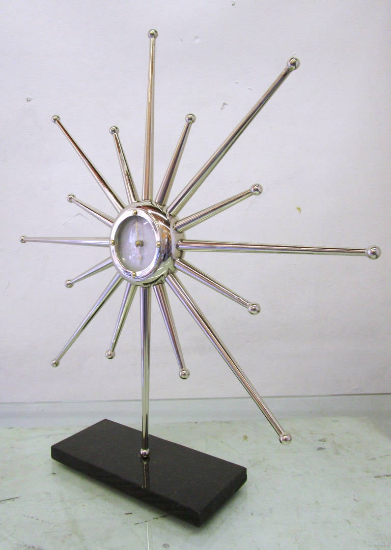 This table clock features a very 1950s starburst design rendered in chrome. The face of the clock is white with brass hands, and is encased by convex glass. The piece is attached to a black stone base via the lowermost ray.