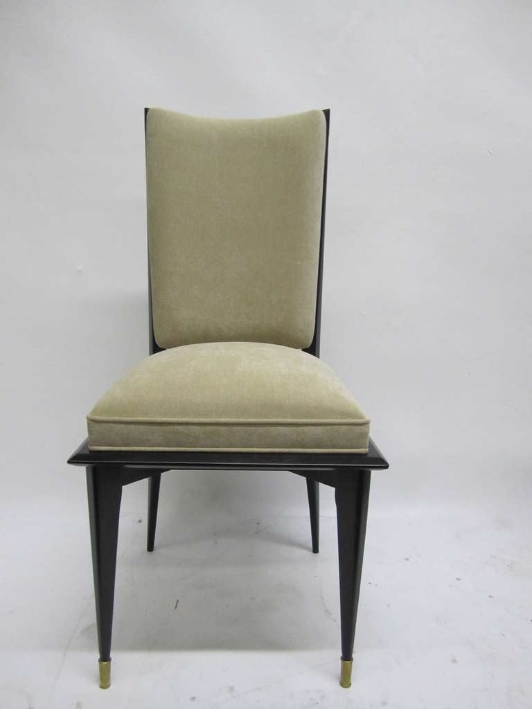 These French, mid-century modern dining chairs feature an elegant, rendered in ebony-stained wood complemented by newly upholstered seat and back rests in cream chenille. The flared, attenuated legs are capped with chrome sabots in front.