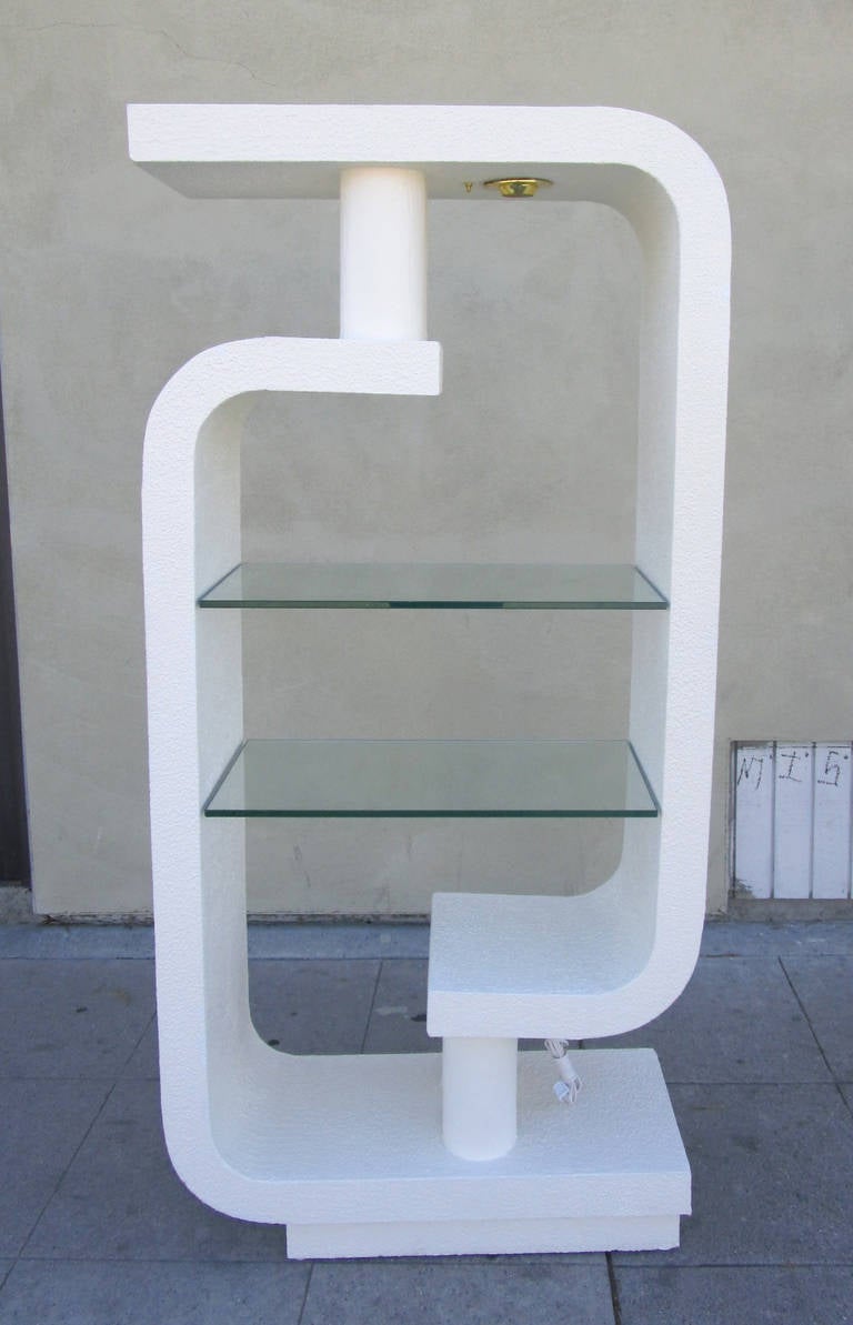 This sculptural shelf  features a curved frame made of two C shaped pieces which are connected via two wood cylinders at the top and bottom. The two glass shelves can be illuminated from above via a built-in light source.
The matching console is