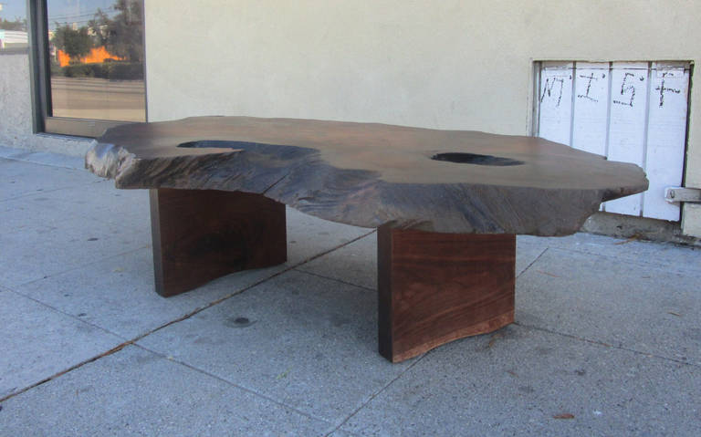 This coffee table after George Nakashima is comprised of a single slab of wood supported by two wide rectangular wooden legs. The amazing natural grain of the wood is enhanced by a cherry-colored stain throughout. This truly unique table is a work