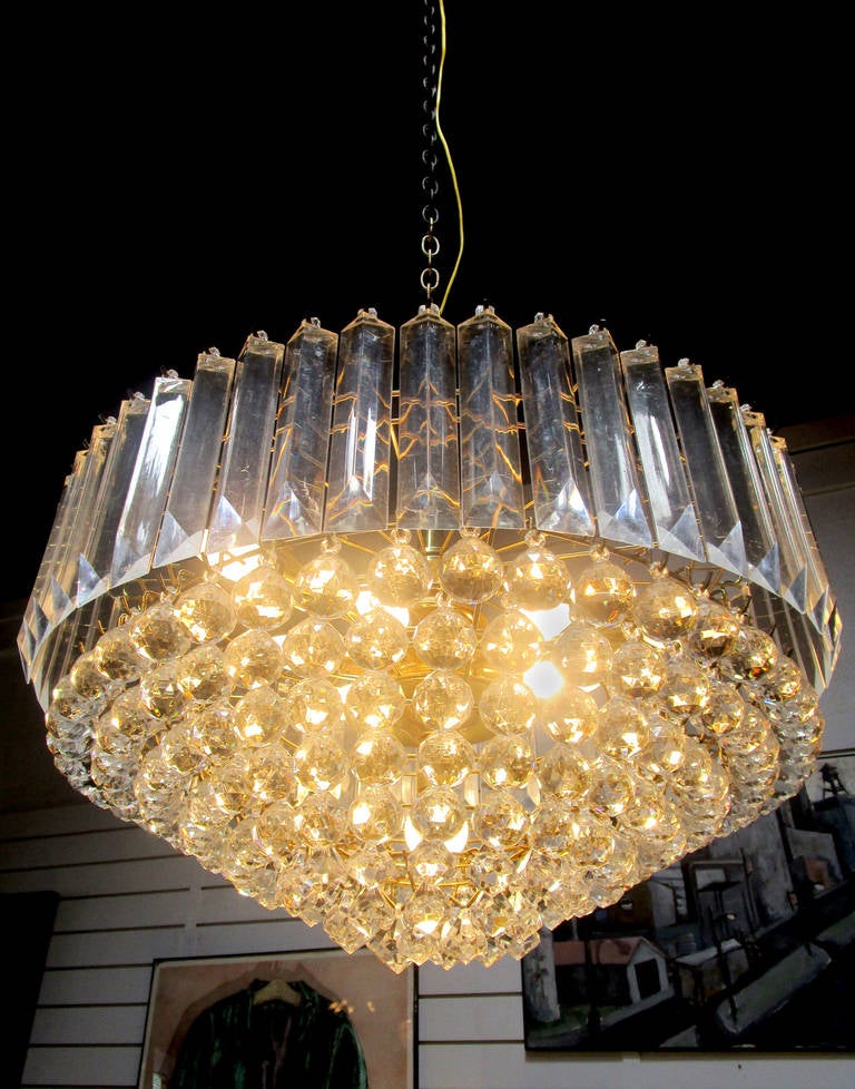 This Lucite chandelier features a wide, slightly tiered brass frame filled in with varying shapes and sizes of Lucite beads. The innermost section has teardrop shaped baubles with slight points at the bottom, followed by a thick ring of faceted